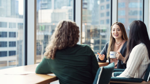 Three people having a conversation at a table in a modern office with city view.